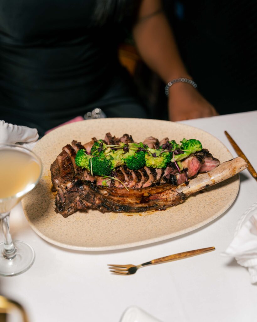 An image of a guest sitting while the waitress drops a plate of steak. The guest has a glass of cocktail on the table.