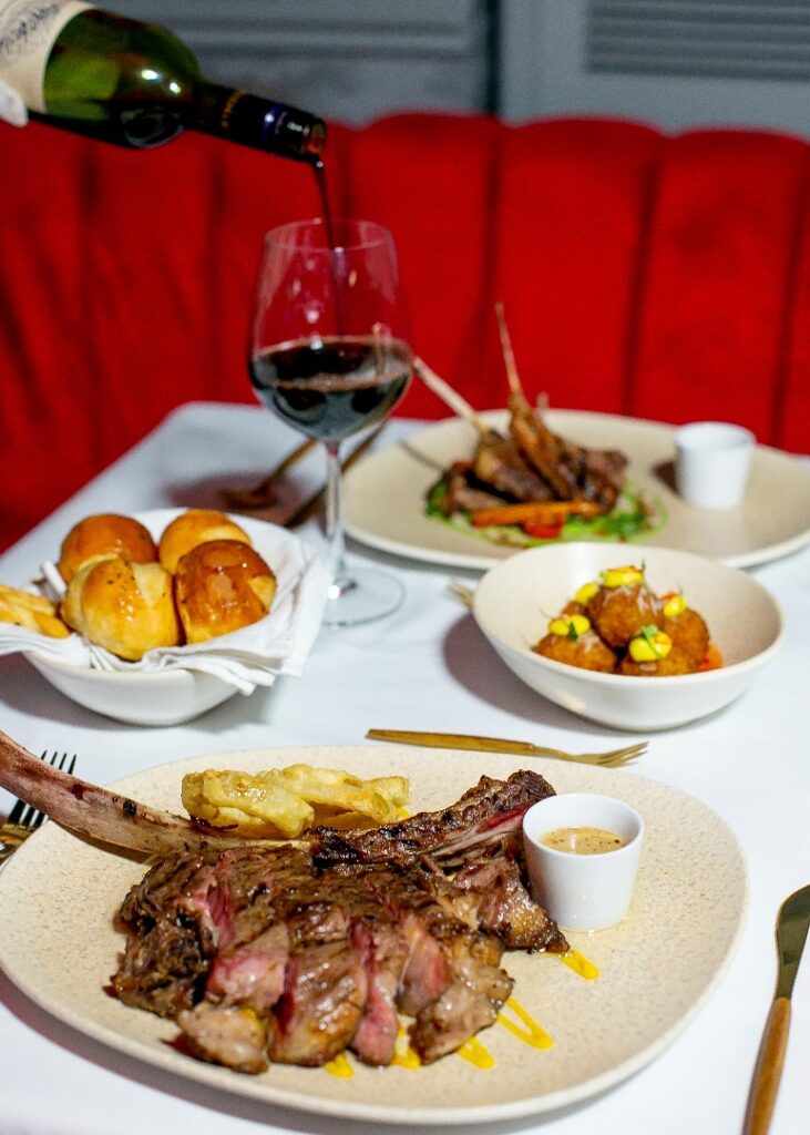 An image of a waitress pouring red wine into a wine glass with the table set. You have two plates of steak and some sides.