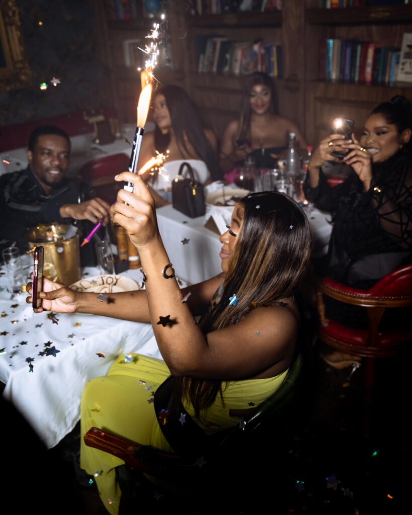 A group of friends at Cee 4 Arena Owerri celebrating a birthday, raising their hands and phones to capture the celebrant as confetti pops and sparklers light up the air, their faces glowing with admiration and excitement